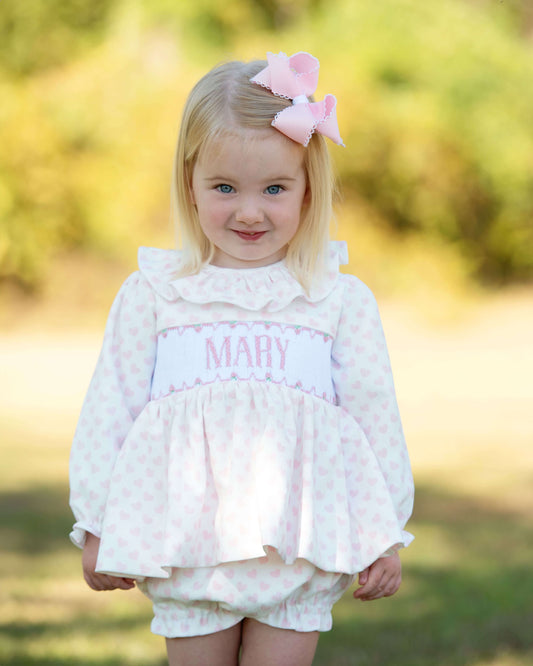 Young girl wearing a pink and white heart outfit with 'MARY' printed on it, standing outdoors.