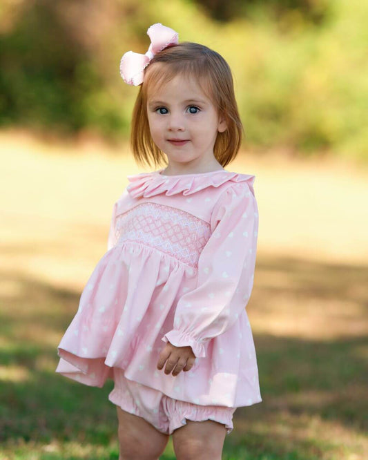 Young girl wearing a pink diaper set with ruffles and a bow in her hair, standing outdoors.