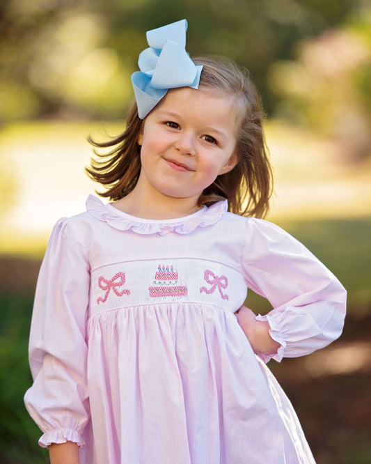 Young girl wearing a pink dress with embroidery and a blue bow in her hair, standing outdoors.