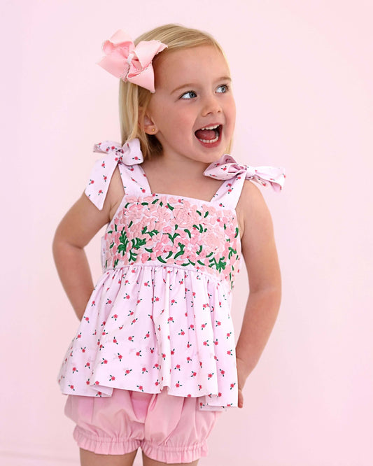 Young girl wearing a pink floral top and shorts with a bow in her hair on a light pink background