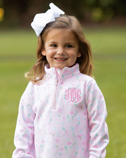 Young girl wearing a pink monogrammed jacket with a white bow in her hair, standing outdoors.