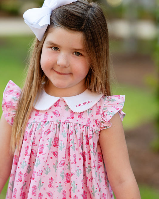 Young girl wearing a pink rodeo dress with a white collar and a large white bow in her hair, standing outdoors.