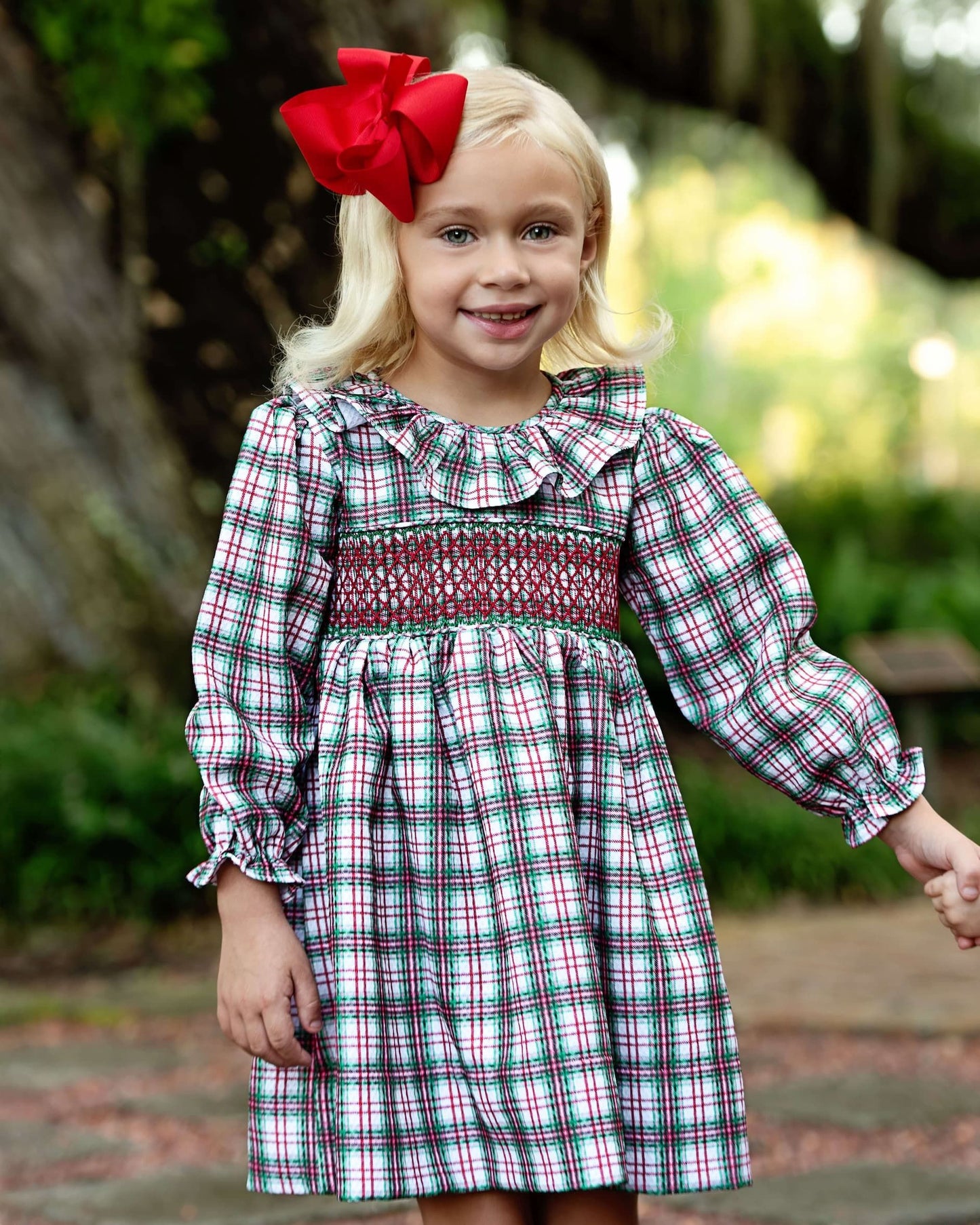 Young girl wearing a plaid dress with a red bow in her hair, standing outdoors.