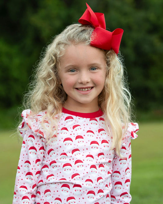 Young girl wearing a red and white patterned outfit with a large red bow in her hair, standing outdoors.