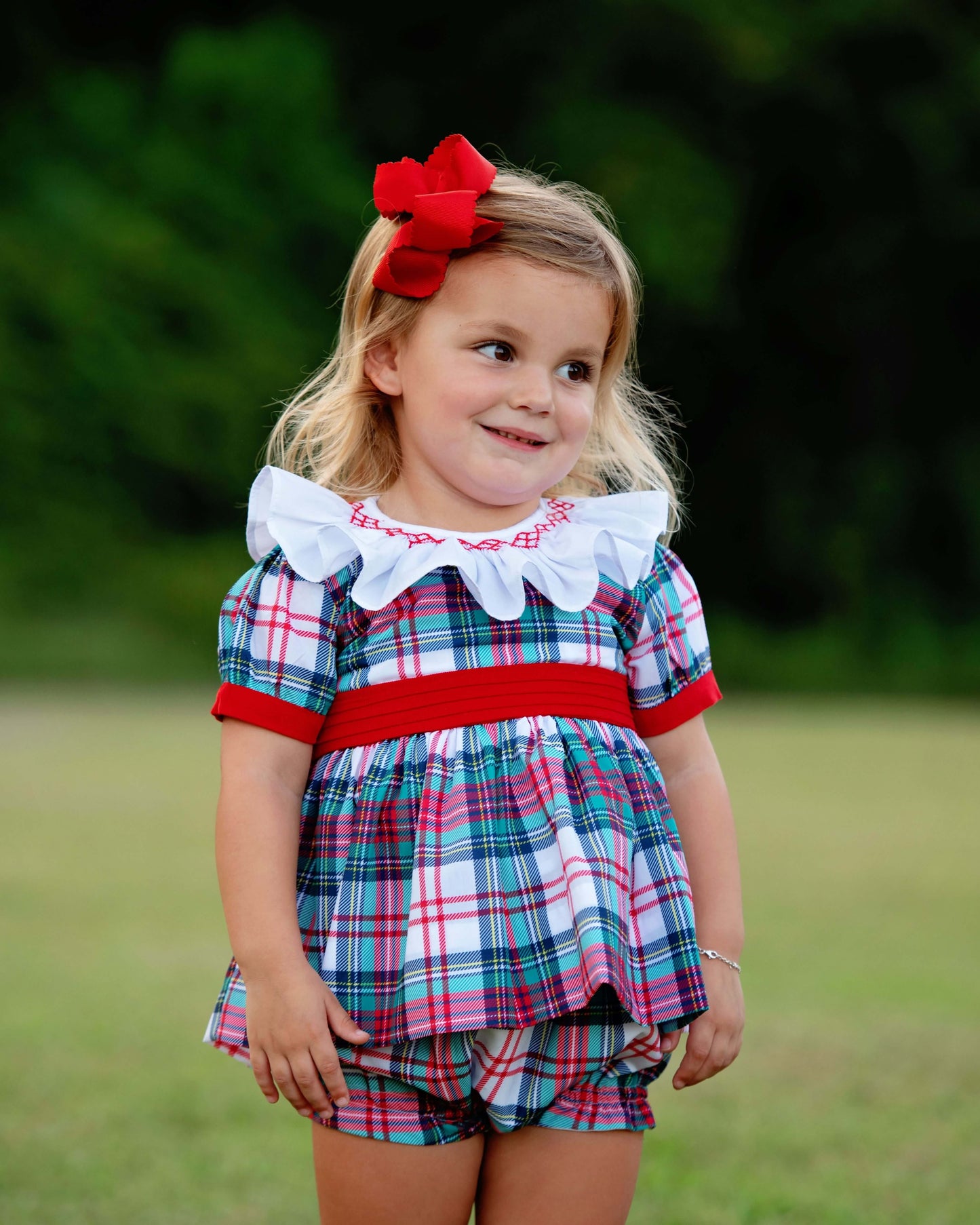 Young girl wearing a smocked plaid diaper set with a red ribbon in her hair, standing outdoors.