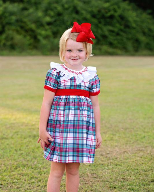 Young girl wearing a smocked plaid dress with a red ribbon in a grassy outdoor setting