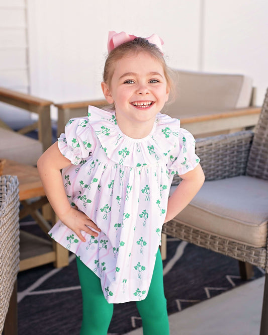 Young girl wearing a white blouse with green floral patterns and green pants, standing outdoors.