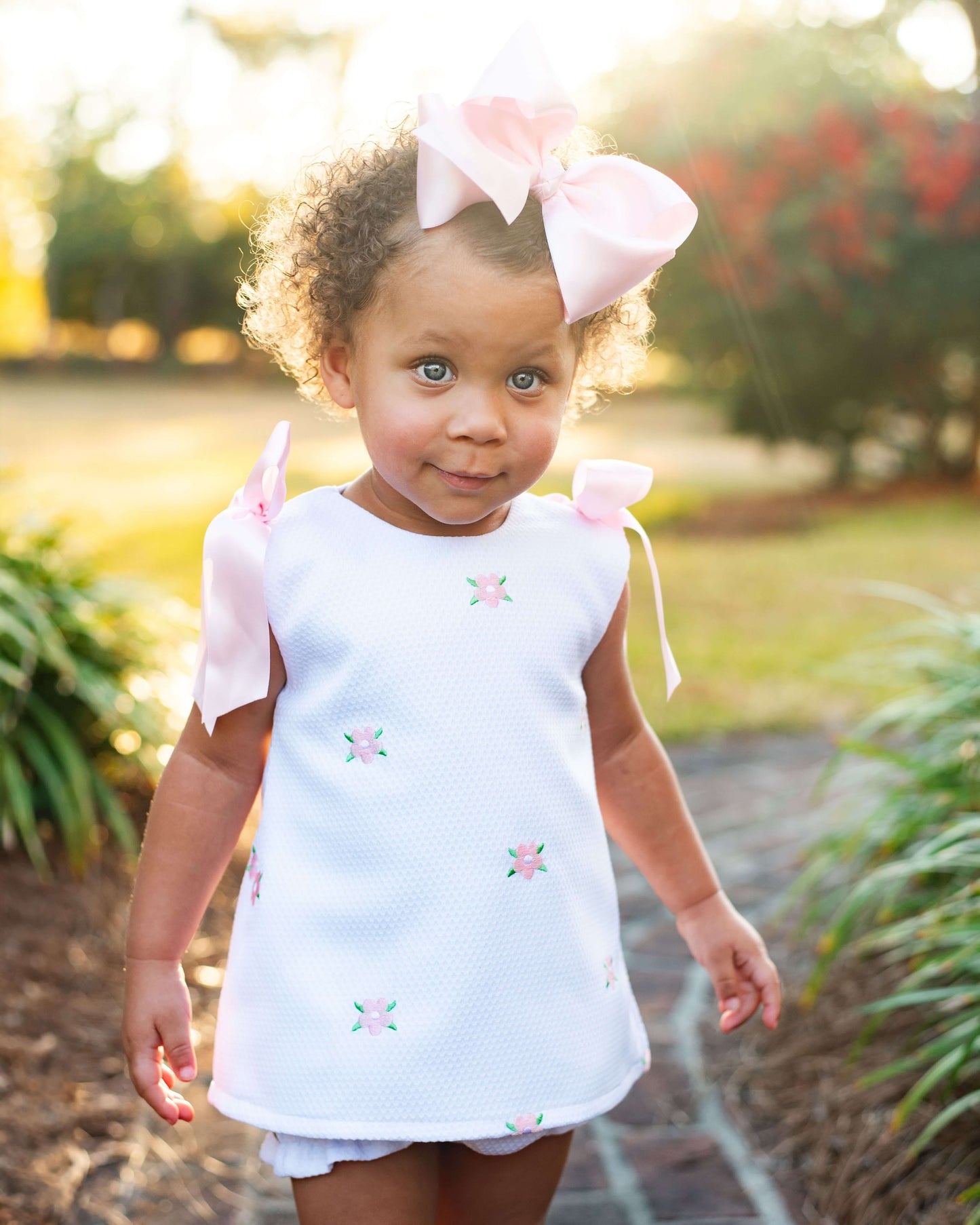 Young girl wearing a white dress embroidered with pink flowers and pink bows on the shoulders, standing outdoors
