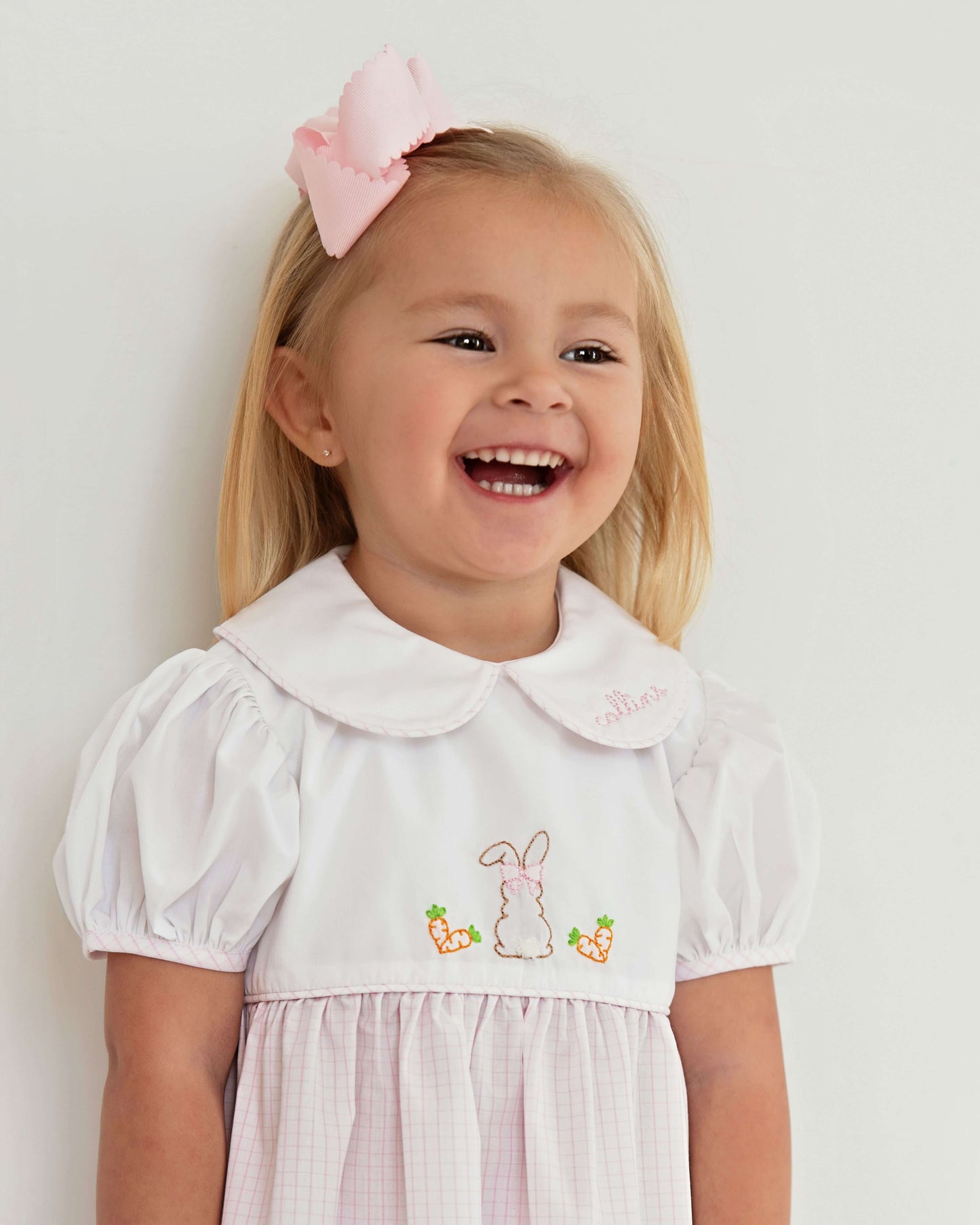 Young girl wearing a white dress with bunny embroidery and a pink bow in her hair against a light background