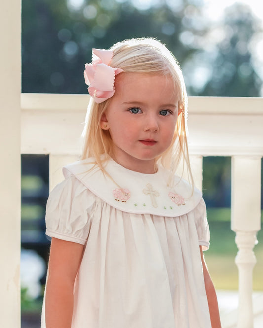 Young girl wearing a white dress with floral embroidery and a pink bow in her hair, standing outdoors.