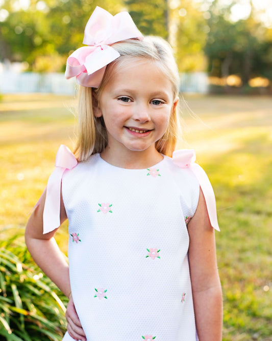 Young girl wearing a white dress with pink bows and floral embroidery in an outdoor setting.