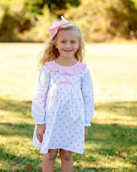 Young girl wearing a white dress with pink hearts and a pink bow outdoors.