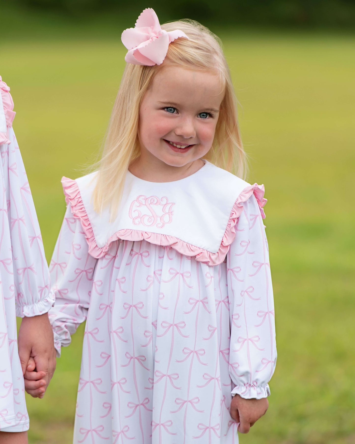 Young girl wearing a white dress with pink ruffles and bow pattern, standing outdoors.