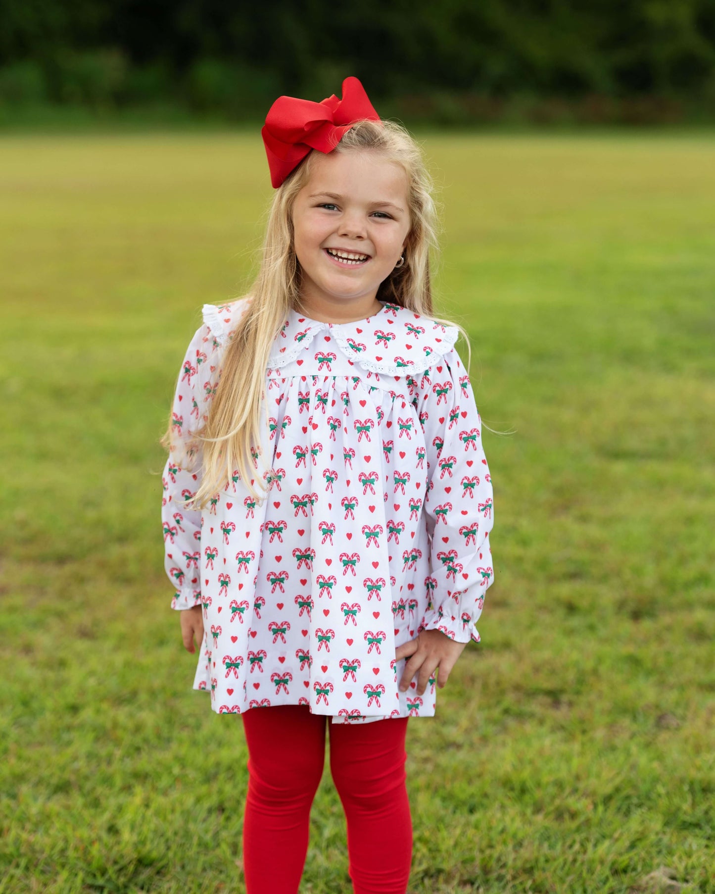 Young girl wearing a white dress with red and green patterns and red leggings standing in a grassy field.