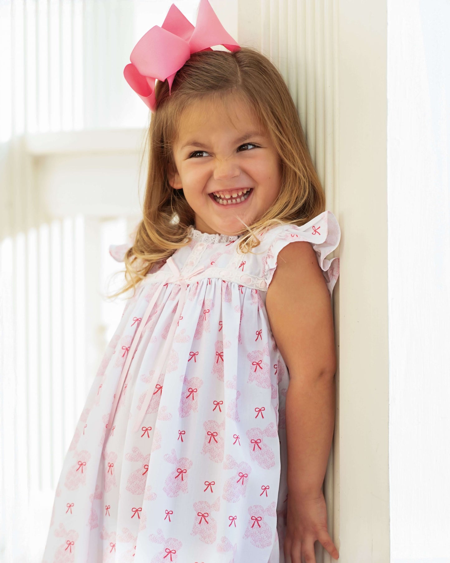 Young girl wearing a white dress with red bows and a pink bow in her hair, standing against a white wall.