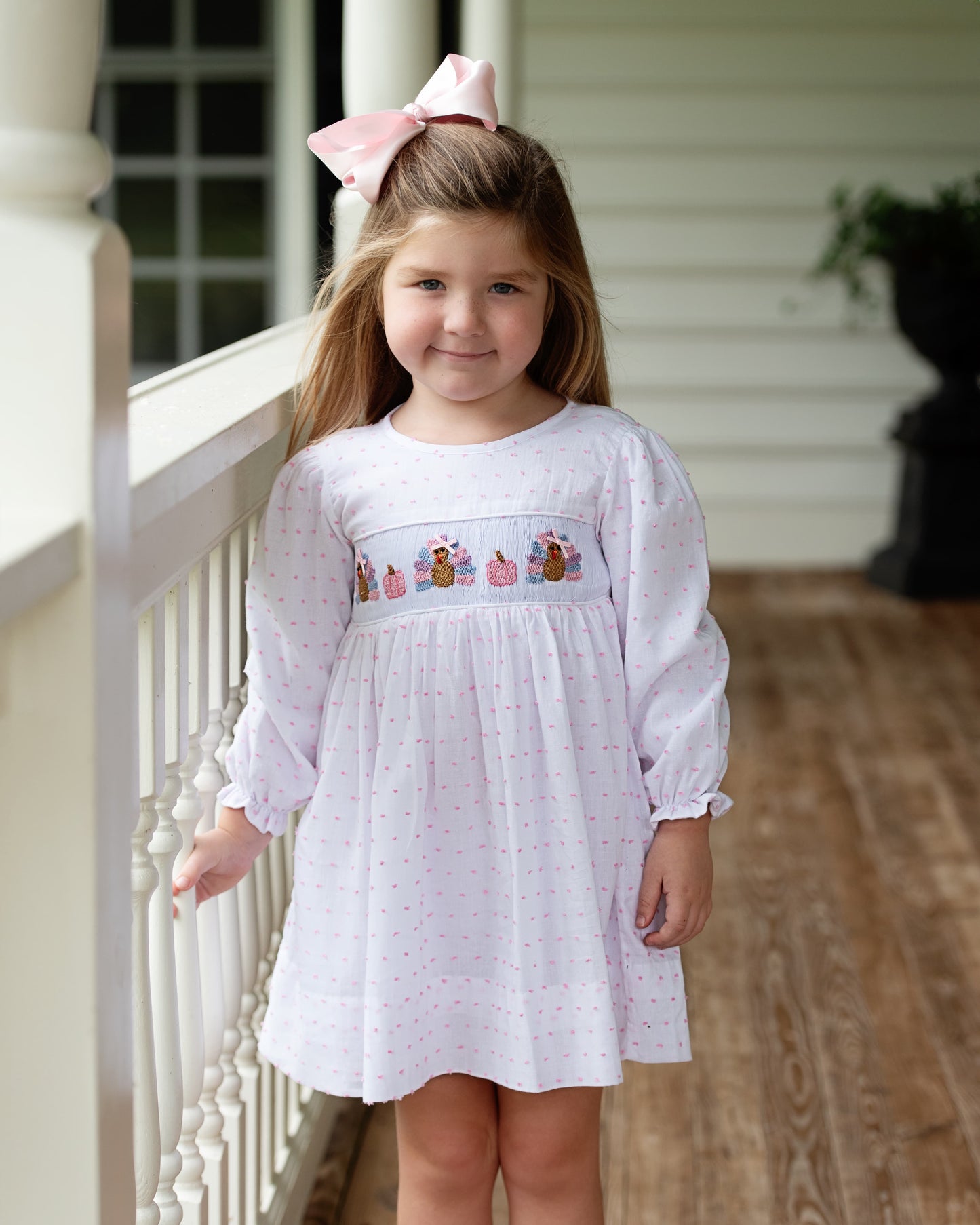 Young girl wearing a white dress with smocked turkeys and pumpkin details on a porch.