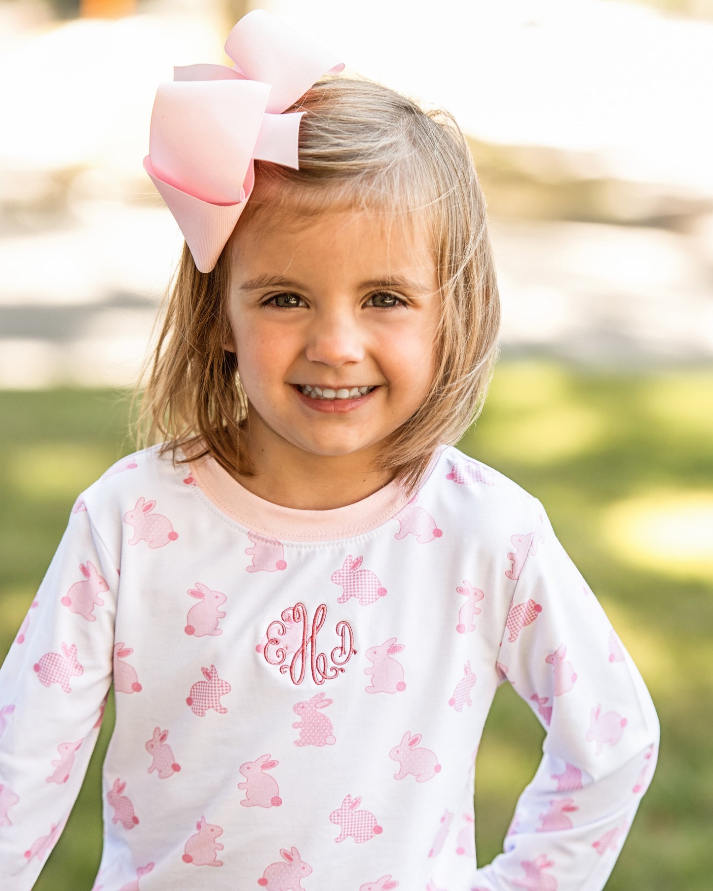 Young girl wearing a white long-sleeve shirt with pink bunny patterns and a large pink bow outdoors.