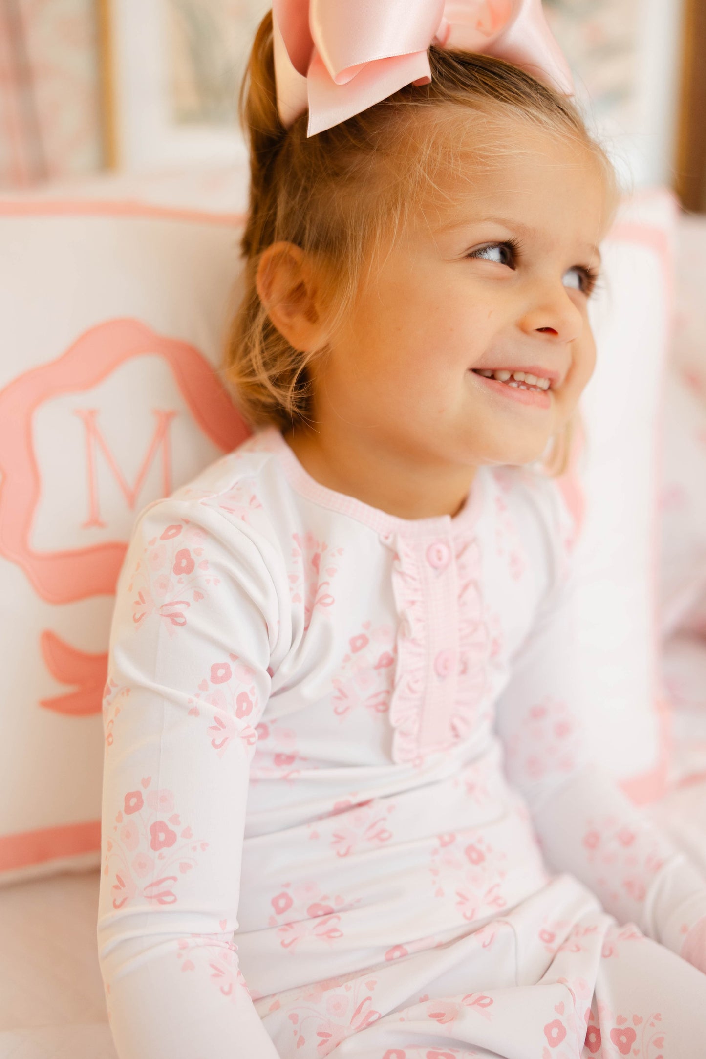 Young girl wearing a white outfit with pink floral patterns, sitting on a pink flamingo cushion.
