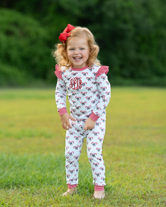 Young girl wearing a white outfit with red bows and monogram, standing in a grassy field.