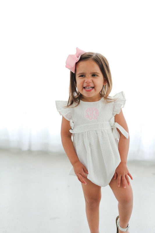 Young girl wearing a white romper with pink text and a pink bow in her hair on a white background