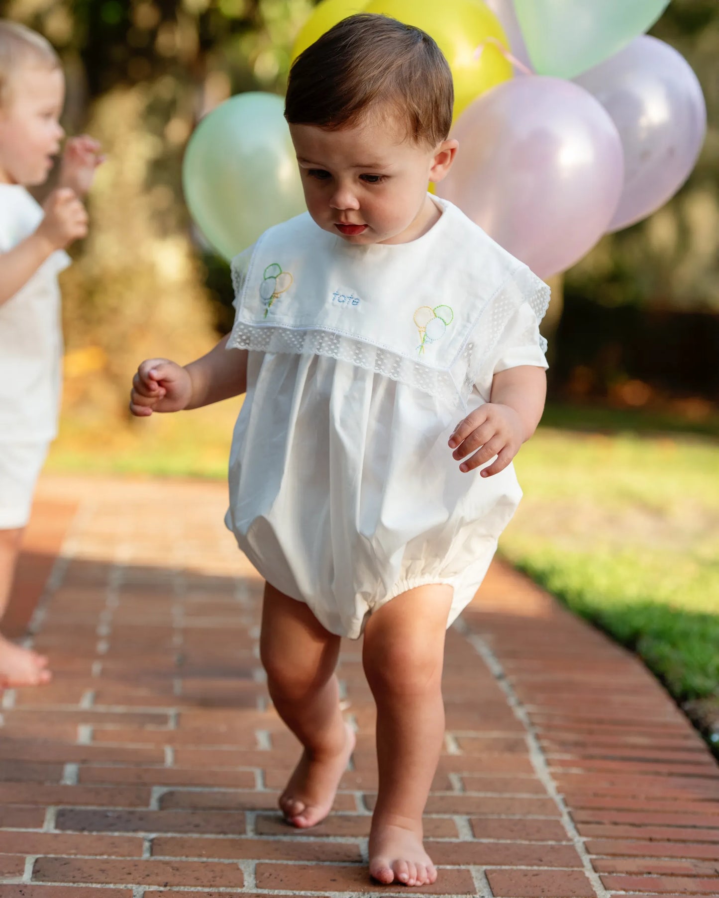 Baby in a white dress walking on a brick path with balloons in the background