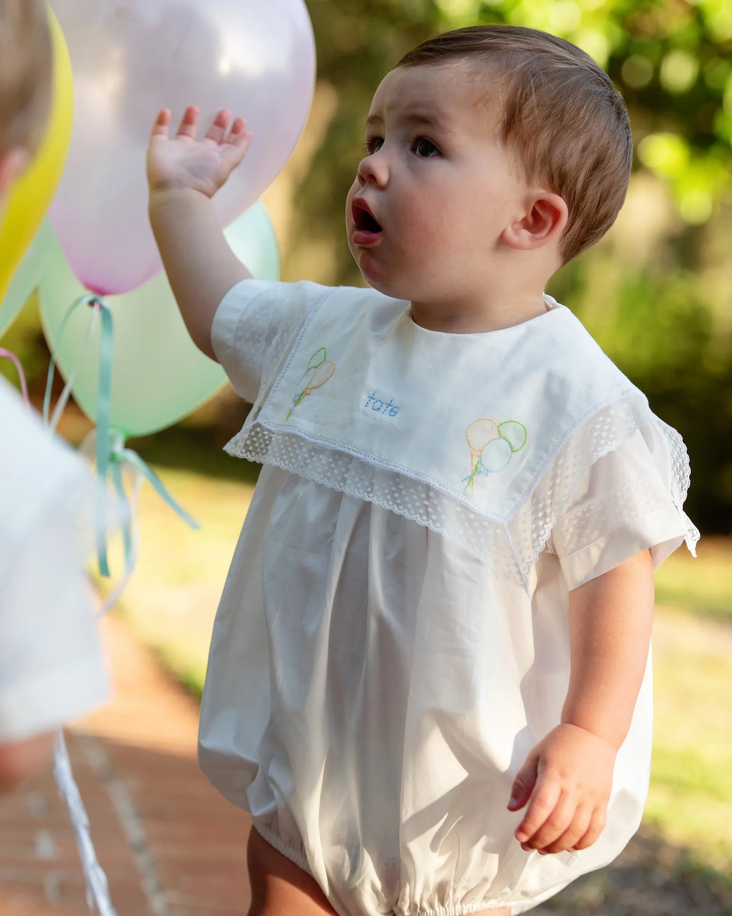 Baby in a white dress with embroidered details, holding balloons outdoors.
