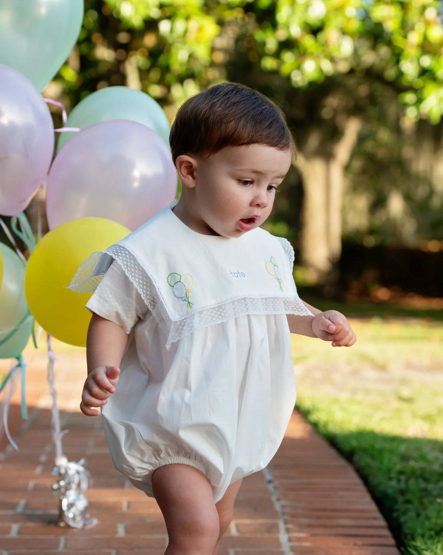 Baby in a white outfit standing outdoors with balloons