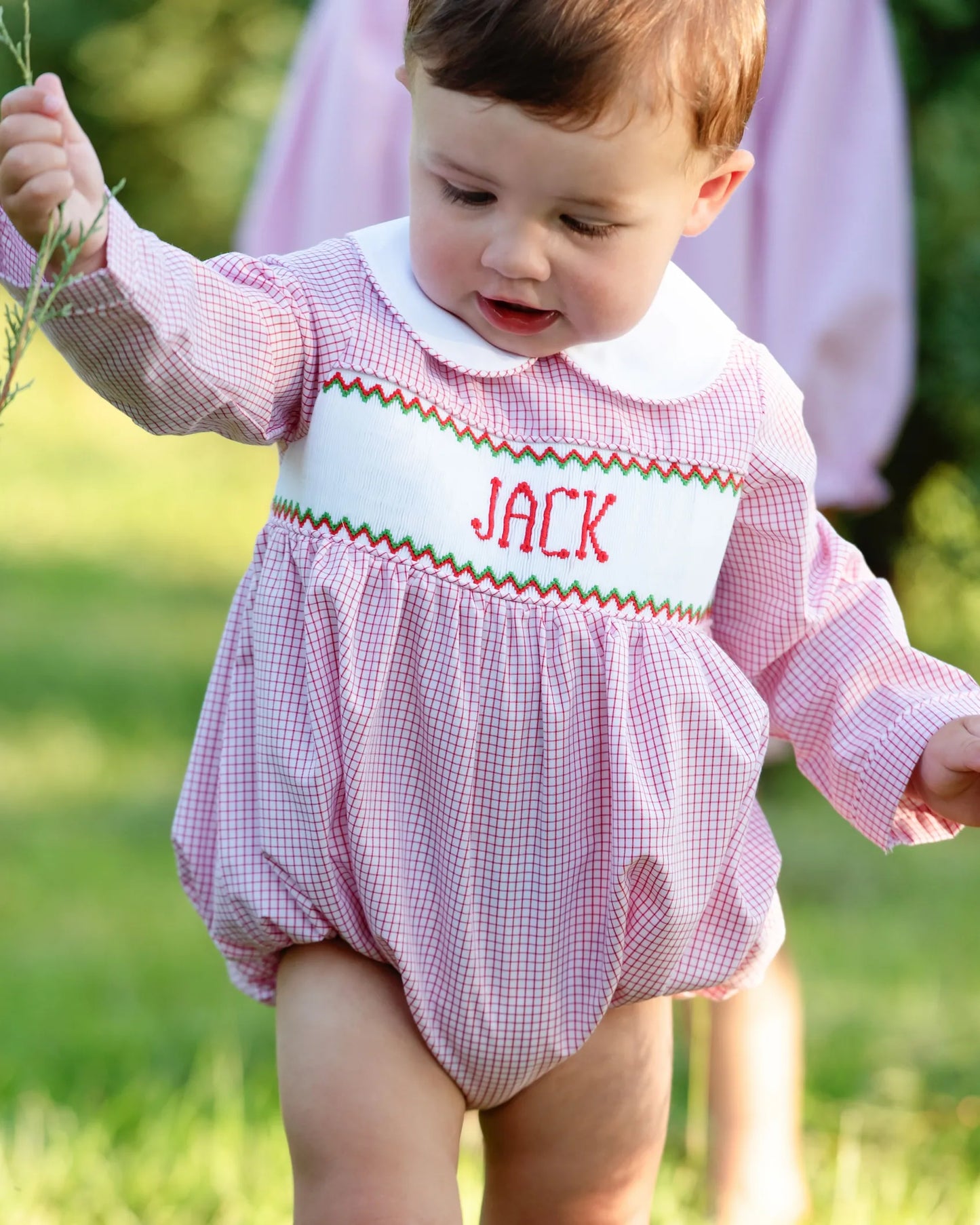 Baby wearing a red checkered romper with 'JACK' printed on it, standing outdoors.
