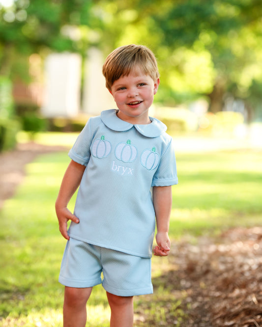 boy wearing a light blue short set with a collar, standing outdoors in a park.