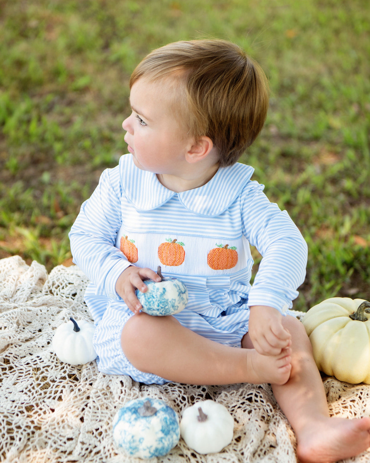 Child in a blue outfit with orange pumpkin patterns sitting on a blanket outdoors.