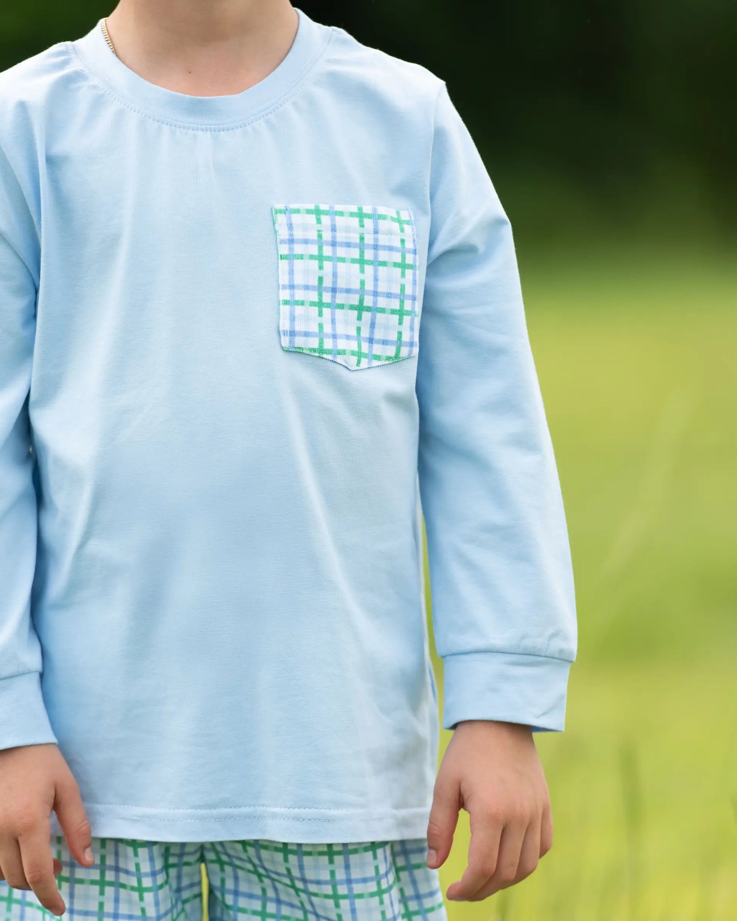 Child wearing a light blue long-sleeve shirt with a green plaid pocket against a blurred natural background.