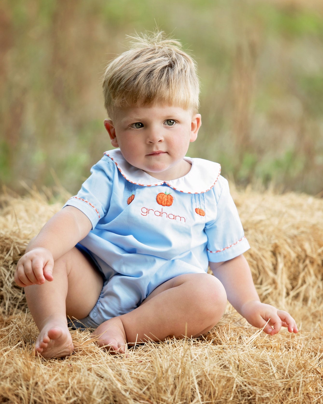 Child wearing a light blue onesie with pumpkin designs sitting on hay.