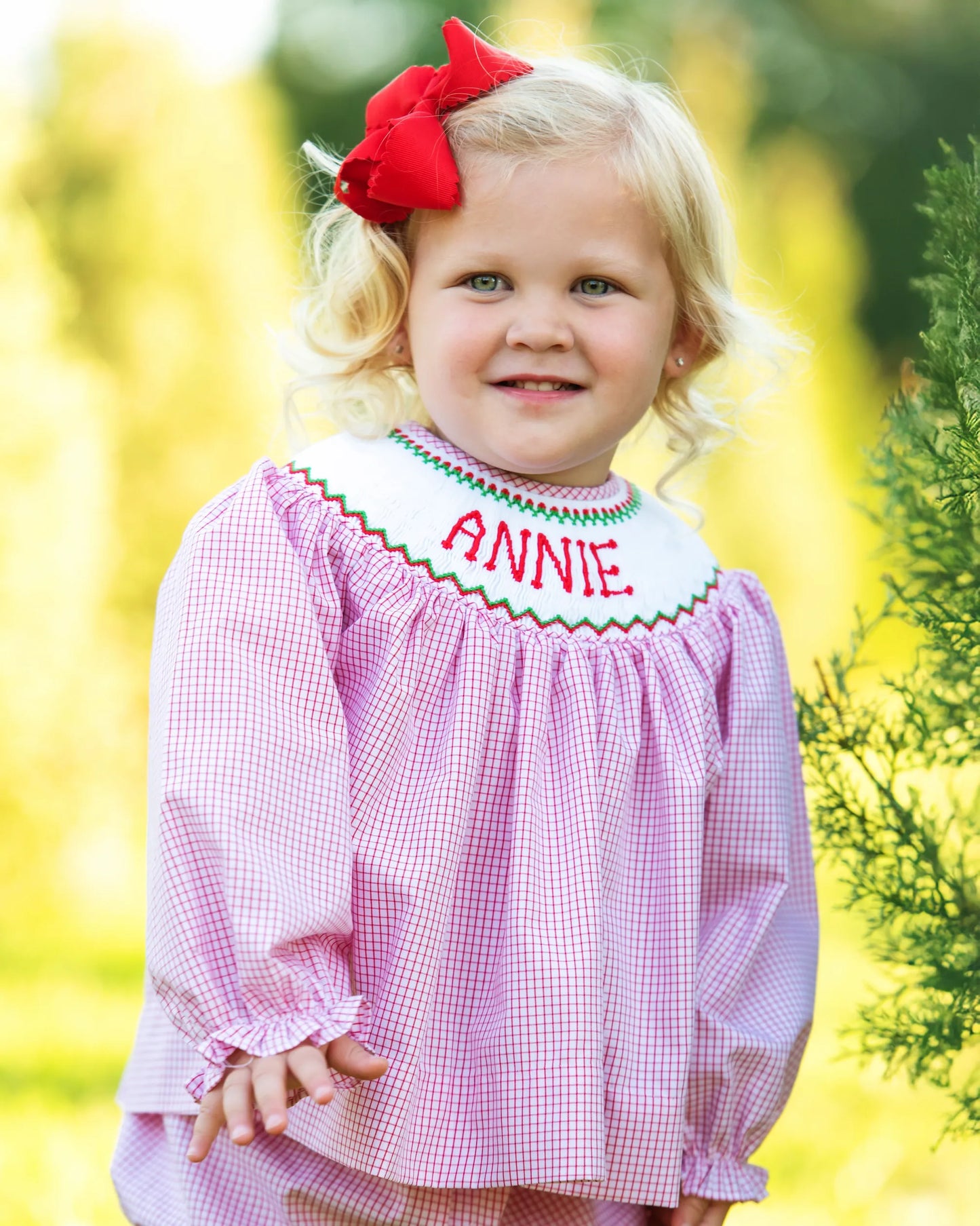 Child wearing a pink checkered dress with 'ANNIE' embroidered on it, outdoors.