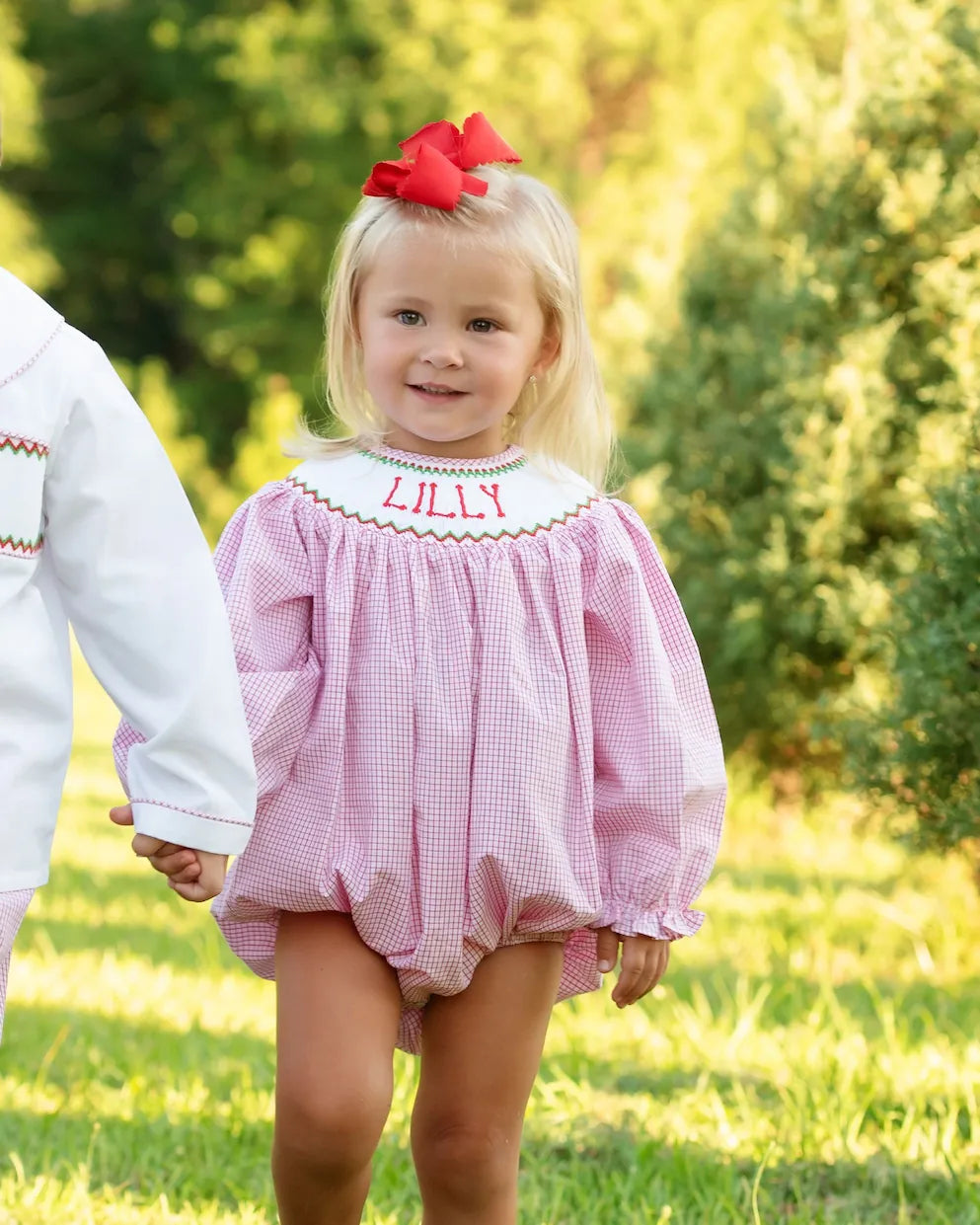 Child wearing a red outfit with 'LILLY' embroidered on it, standing outdoors.