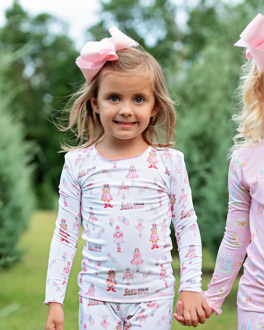 A young girl wearing a white long-sleeve pajama top and matching pants with a pink and red ballerina print, accessorized with a large pink bow, standing outdoors and holding hands with another child.