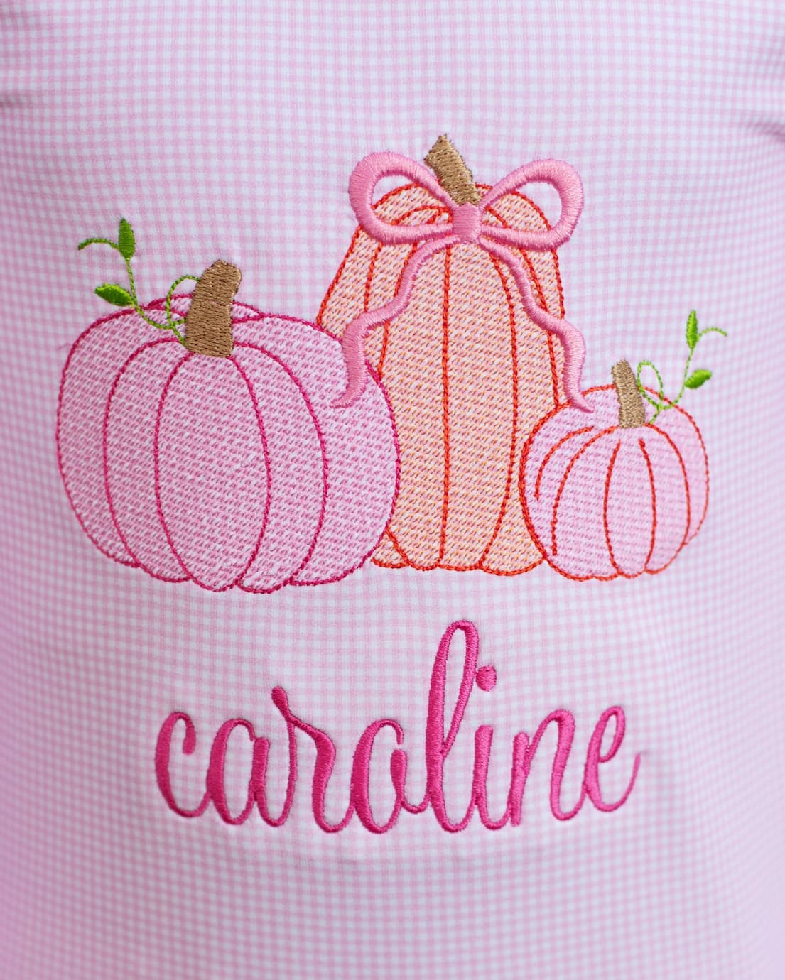 Embroidered design of pumpkins with a pink bow and the name 'caroline' on a light pink background.