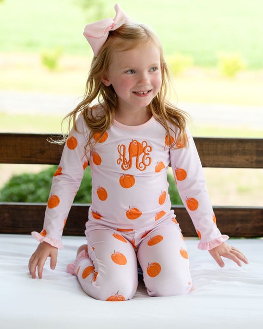 girl wearing a pink pajama set with orange pumpkin patterns and pink monogram "aAe", sitting outdoors.