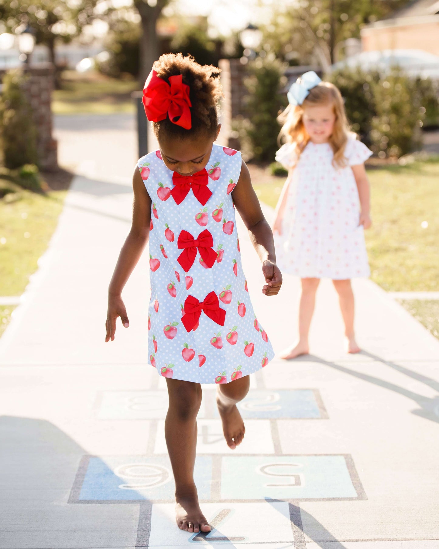 girl wearing apple print dress with with red bows down the front hopping on one leg.