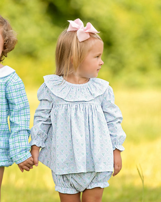 Two children holding hands in a field with one wearing a light blue dress and pink bow.