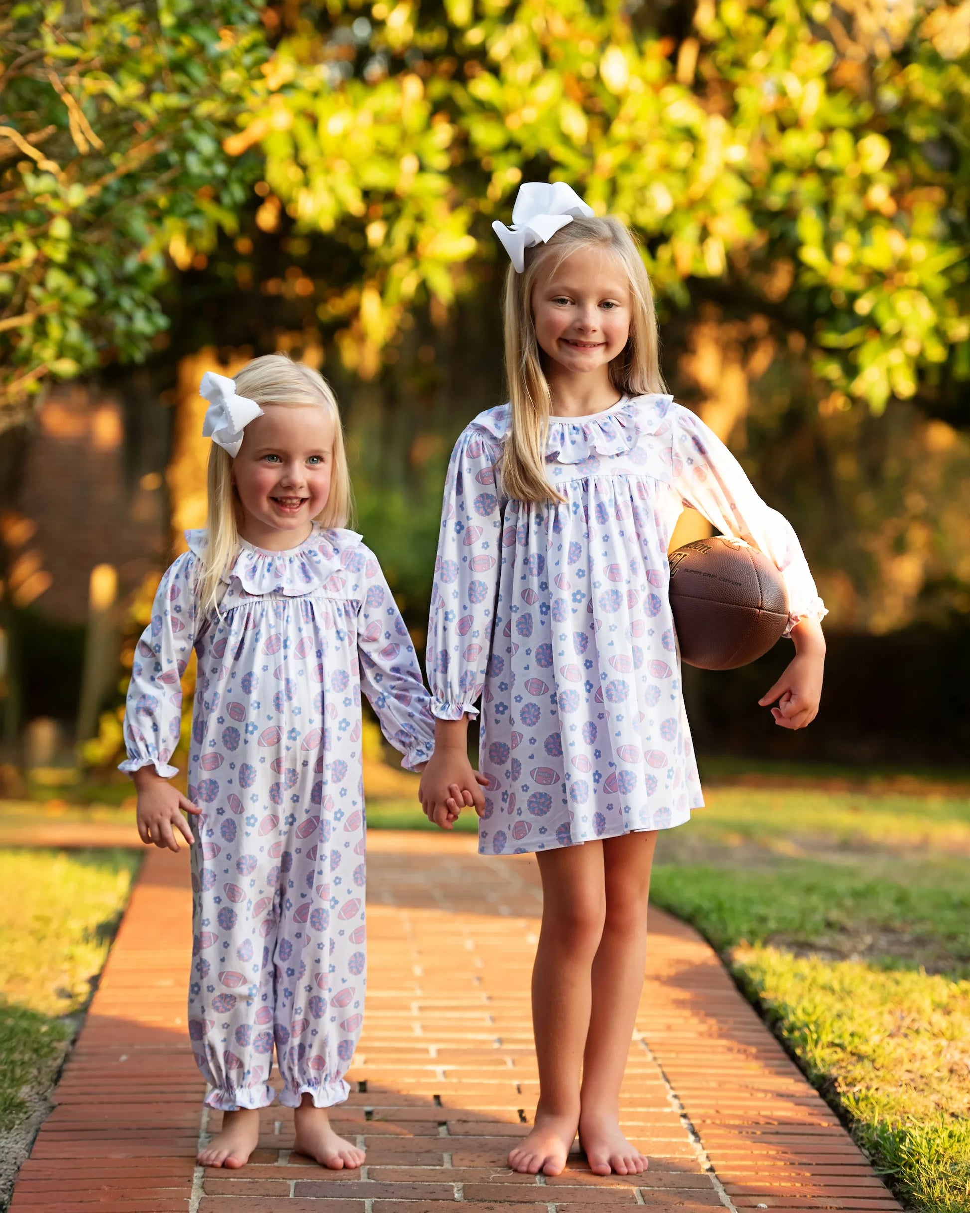 Two young girls in matching dresses holding hands on a brick path with greenery in the background.