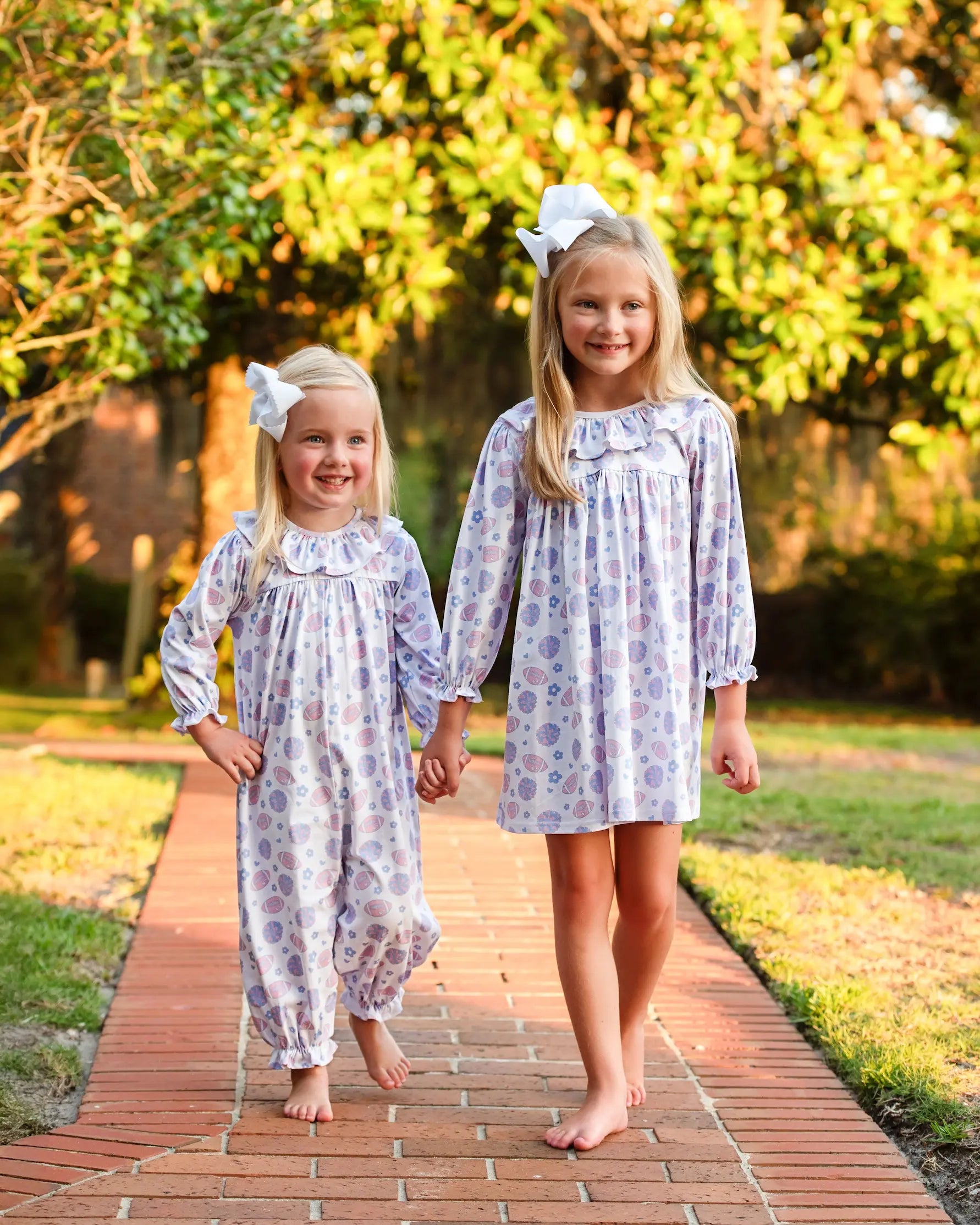 Two young girls in matching dresses standing on a brick path with trees in the background.