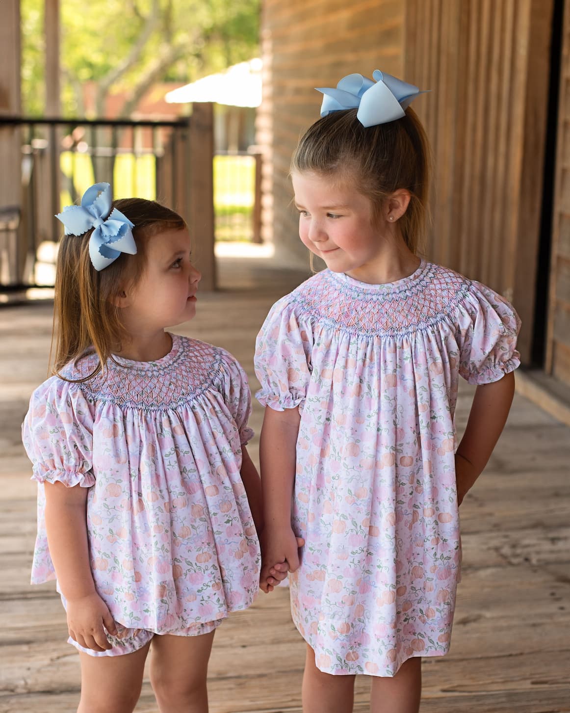 Two young girls wearing matching floral dresses with large blue bows outdoors.
