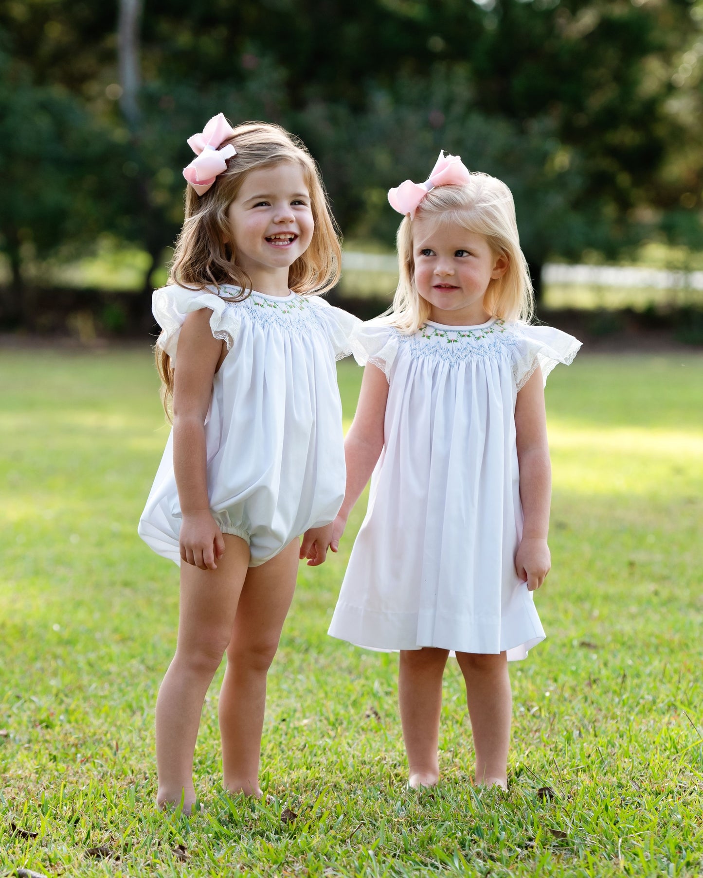 two small girls wearing matching smocked outfits, standing outside with pink bows in their hair