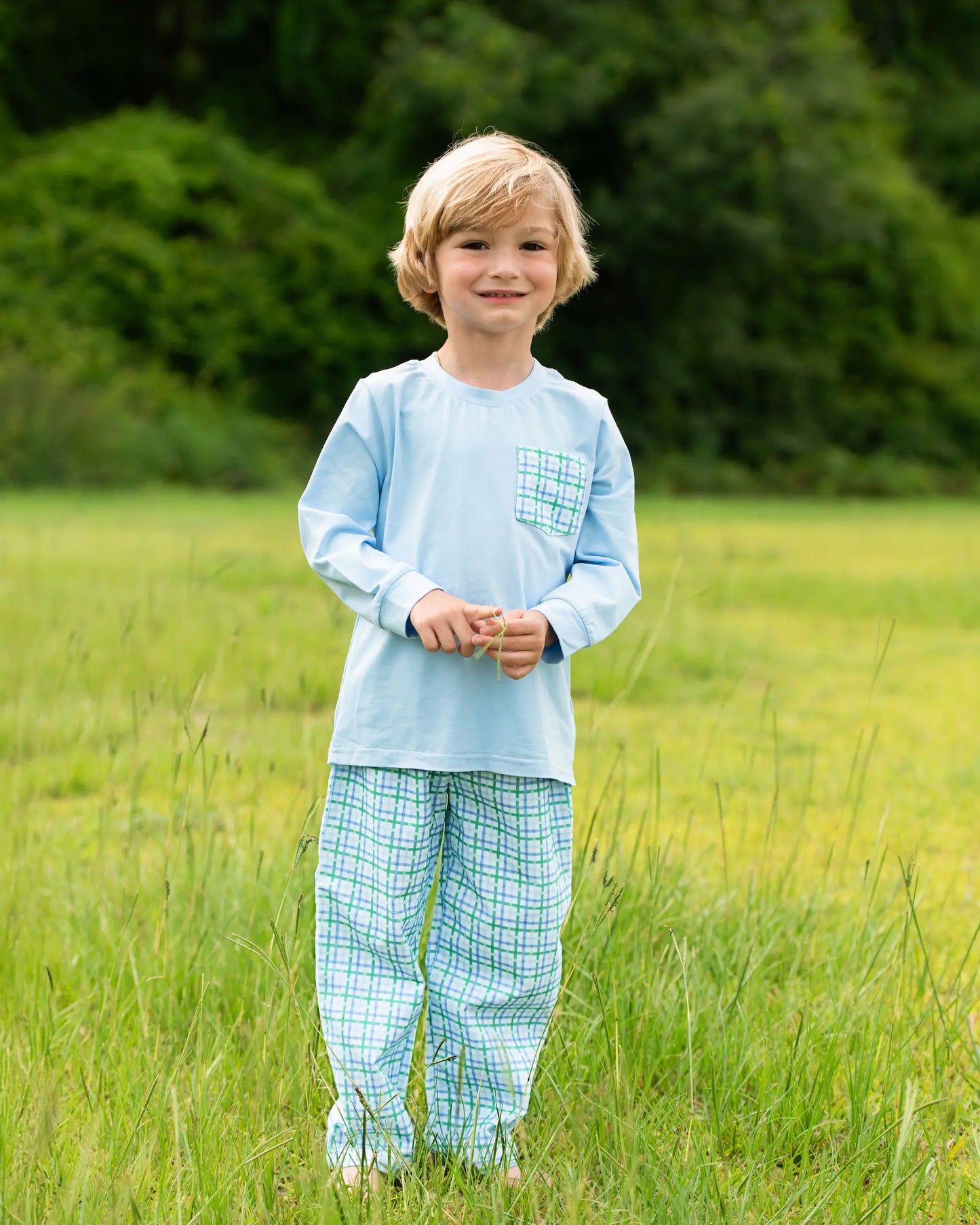Young boy in a light blue shirt and green checkered pants standing in a grassy field.