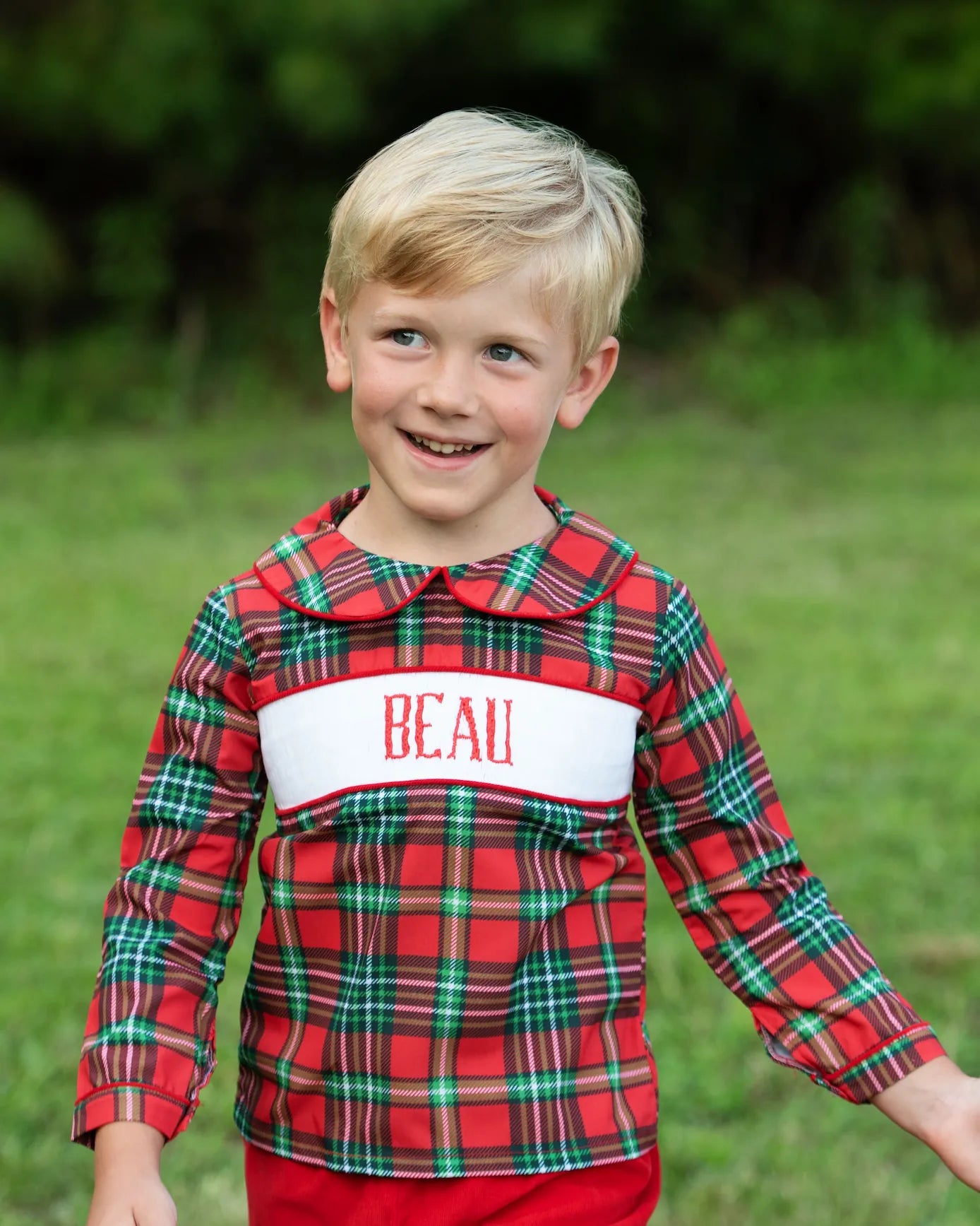 Young boy wearing a red plaid shirt with 'BEAU' on it, standing outdoors.