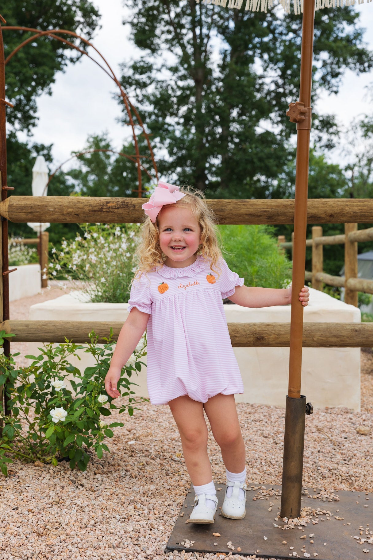Young girl in a pink dress standing outdoors with greenery and wooden structure in the background