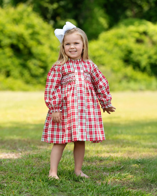 Young girl in a red and white checkered dress standing outdoors with greenery in the background