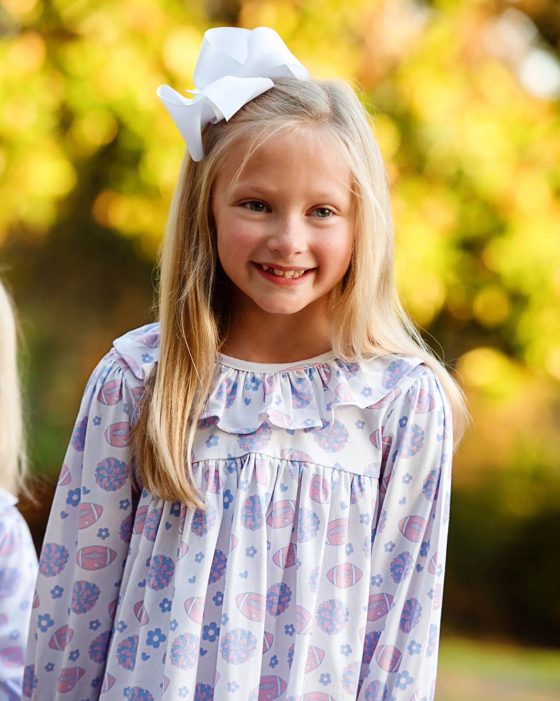 Young girl wearing a floral dress with a white bow in her hair, standing outdoors with a blurred natural background.