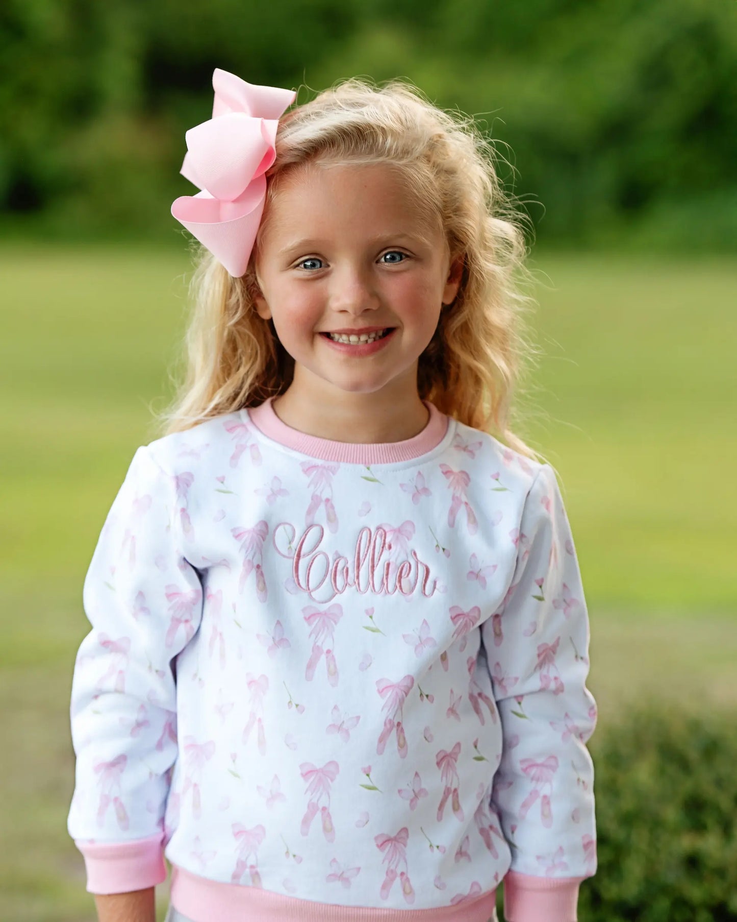 Young girl wearing a light blue sweatshirt with pink bows and the name 'Collier' on it, standing outdoors.