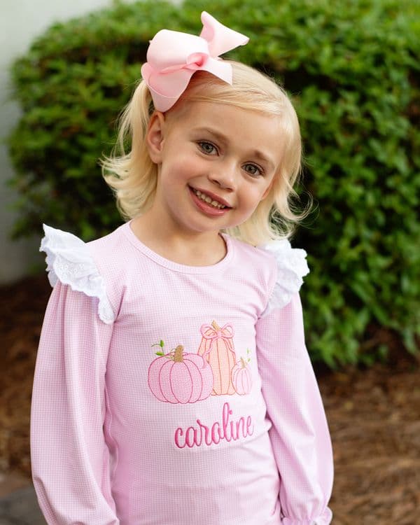 Young girl wearing a pink outfit with pumpkins and the name 'Caroline' outdoors.